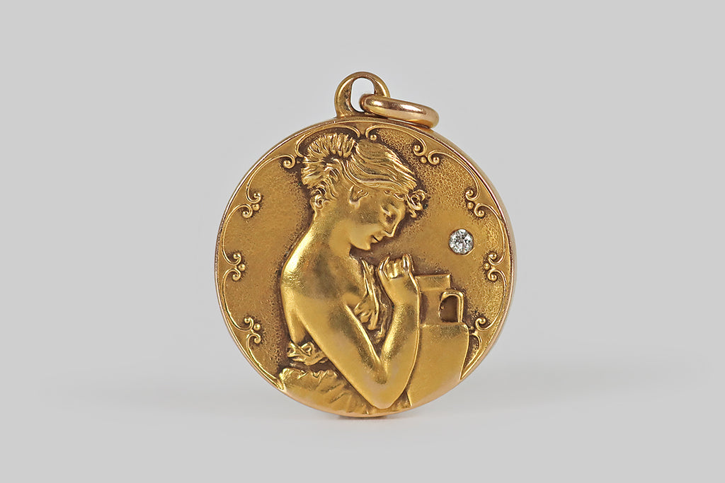 Poor Mouchette | Curated Antique Jewelry, Vintage Jewelry & Engagement Rings | Portland, Oregon | An Art Nouveau era figural locket, modeled in 14k yellow gold,&nbsp;whose decor features a woman in profile, dressed in a grecian gown. This lady wears a gentle&nbsp;expression; her hair is bound up in a French twist; she&nbsp;gazes thoughtfully at the urn&nbsp;sitting beside her. This scene is surmounted by a stylized, scalloped border. A small, sparkling old mine cut diamond
