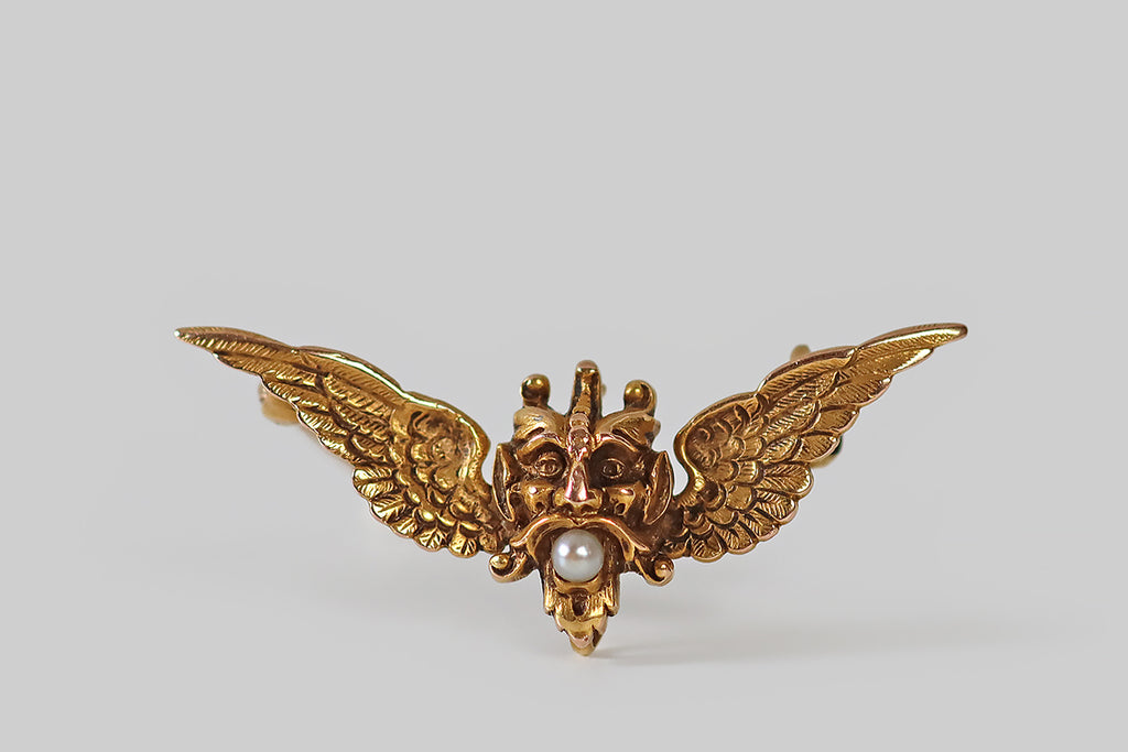 Antique Jewelry Portland, Vintage Jewelry Portland , Antique Engagement Rings | Poor Mouchette | A wonderful, small, Art Nouveau era brooch, modeled in 14k yellow gold, as the head of a devil, flanked by a pair of feathered wings. Our impish friend holds a small, natural, white sea pearl in his mouth, He wears a pointy, van dyke beard and mustache, along with a lively expression. His ears are pointy, and horns protrude from his head. We see Lucifer, the fallen angel, in this depiction