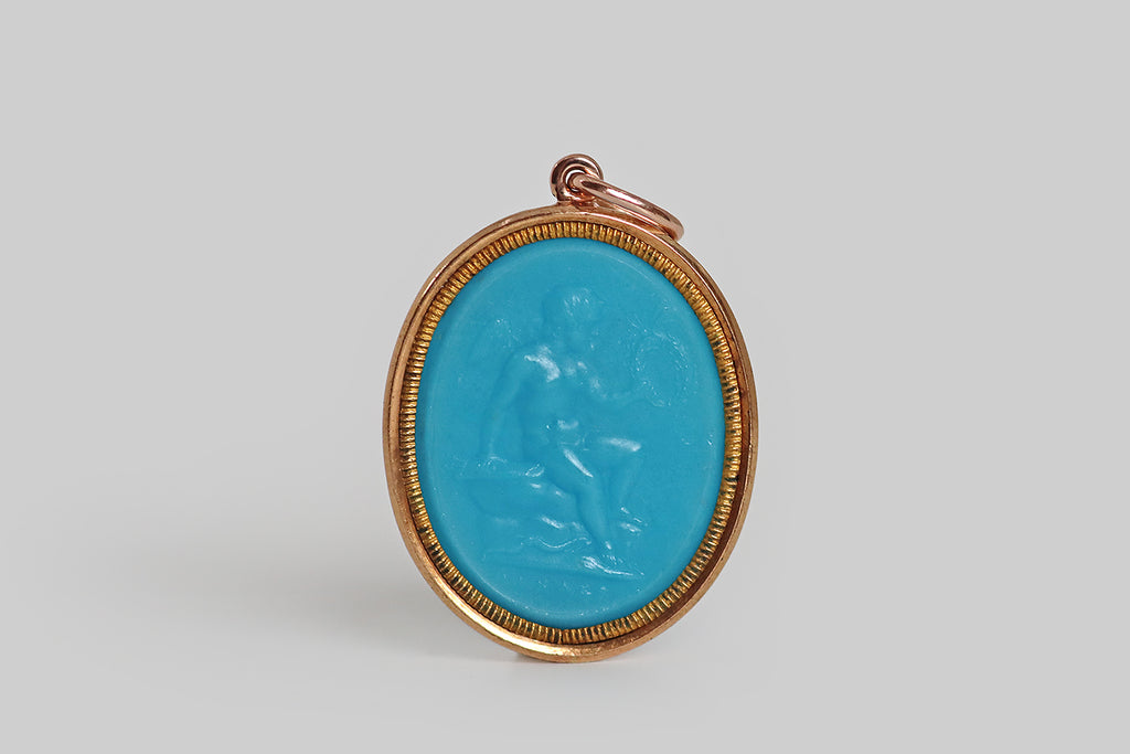 Antique Jewelry Portland, Vintage Jewelry Portland , Antique Engagement Rings | Poor Mouchette | A wonderful Georgian-era pendant, modeled in 18k yellow gold, whose primary element is a rare, opaque, turquoise-blue, glass cameo. This cameo depicts cupid (Eros), resting on a stone and holding aloft a wreath of laurels. The cameo is bordered by an inner bezel, and this bezel is decorated with fine notches that give it a coiled appearance.