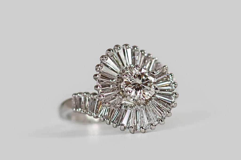 Antique Jewelry Portland, Vintage Jewelry Portland , Antique Engagement Rings | Poor Mouchette | A striking, vintage, ballerina-style cluster ring, modeled in platinum, whose primary gem is a .65 carat round brilliant cut diamond (G/H, VS). This vibrant, white diamond is held in six prongs, at the center of the ring face. Surrounding it, two curving rows of tapered baguette diamonds create the impression of a spiral, or a double helix, moving around the central stone.