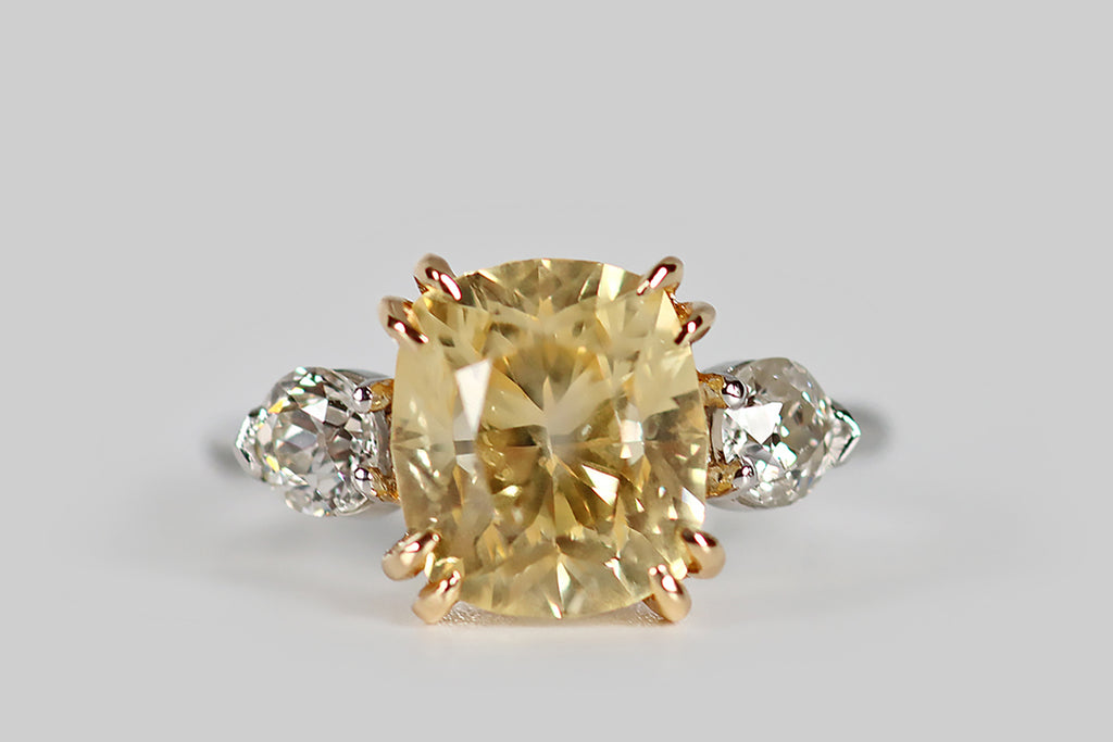 Poor Mouchette | Curated Antique Jewelry, Vintage Jewelry & Engagement Rings | An elegant three-stone engagement ring, modeled in platinum and 18k yellow gold, whose primary gem is a natural, antique, 5.7 carat, cushion-cut yellow sapphire. This pillowy sapphire has a soft, icy-yellow color— it glitters cheerfully atop the ring face, where it rests in a yellow gold basket. This sapphire is held in place by four pairs of tapering, claw-like, yellow gold prongs.