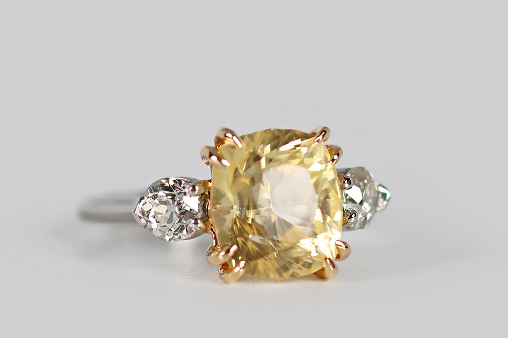 Poor Mouchette | Curated Antique Jewelry, Vintage Jewelry & Engagement Rings | An elegant three-stone engagement ring, modeled in platinum and 18k yellow gold, whose primary gem is a natural, antique, 5.7 carat, cushion-cut yellow sapphire. This pillowy sapphire has a soft, icy-yellow color— it glitters cheerfully atop the ring face, where it rests in a yellow gold basket. This sapphire is held in place by four pairs of tapering, claw-like, yellow gold prongs.