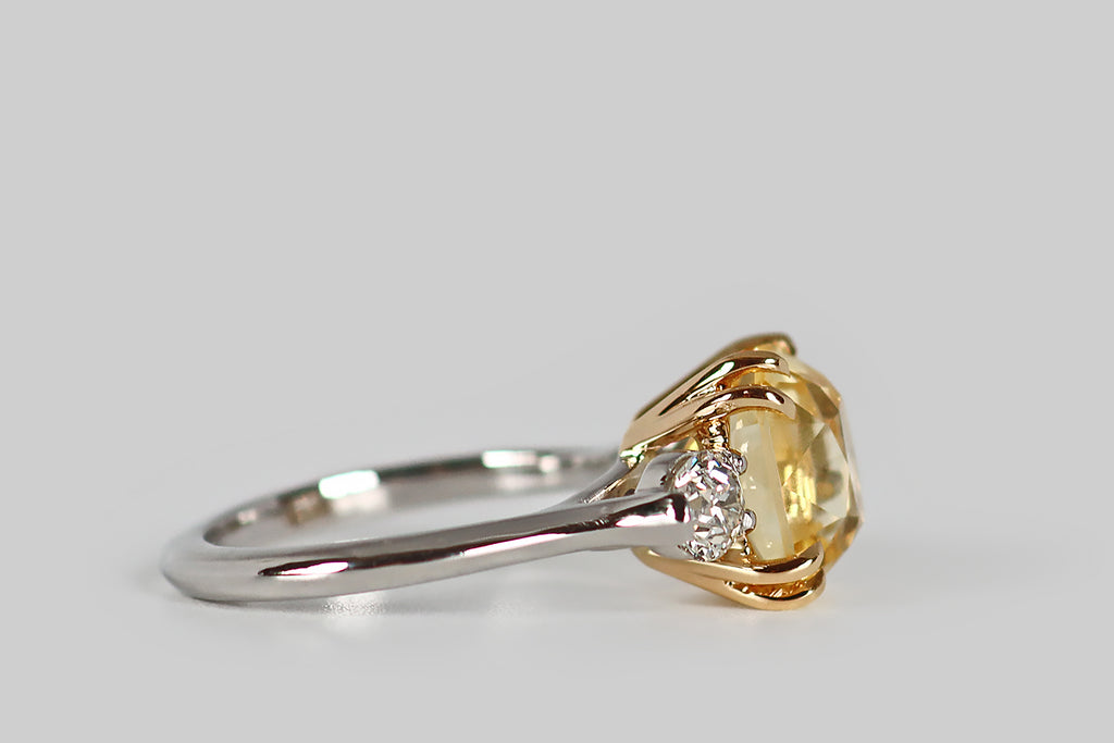 Poor Mouchette | Curated Antique Jewelry, Vintage Jewelry & Engagement Rings | An elegant three-stone engagement ring, modeled in platinum and 18k yellow gold, whose primary gem is a natural, antique, 5.7 carat, cushion-cut yellow sapphire. This pillowy sapphire has a soft, icy-yellow color— it glitters cheerfully atop the ring face, where it rests in a yellow gold basket. This sapphire is held in place by four pairs of tapering, claw-like, yellow gold prongs.
