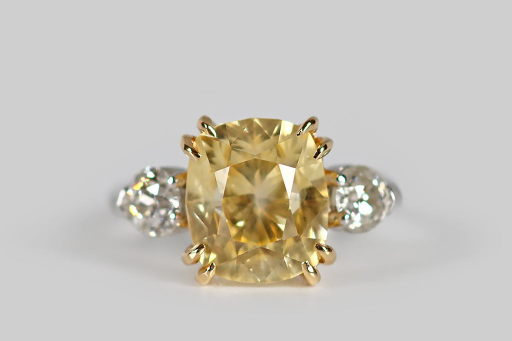 Poor Mouchette | Curated Antique Jewelry, Vintage Jewelry & Engagement Rings | An elegant three-stone engagement ring, modeled in platinum and 18k yellow gold, whose primary gem is a natural, antique, 5.7 carat, cushion-cut yellow sapphire. This pillowy sapphire has a soft, icy-yellow color— it glitters cheerfully atop the ring face, where it rests in a yellow gold basket. This sapphire is held in place by four pairs of tapering, claw-like, yellow gold prongs.