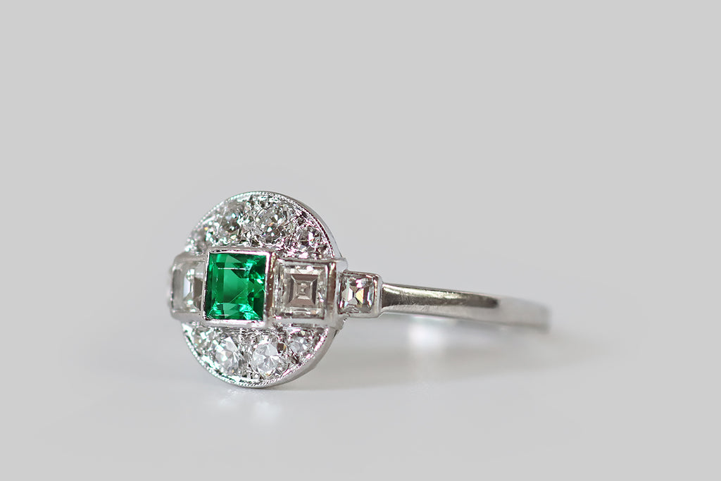 Antique Jewelry Portland, Vintage Jewelry Portland , Antique Engagement Rings | Poor Mouchette | An unusual Art Deco era engagement ring, modeled in platinum, whose primary gem is a vibrant, highly-saturated, step cut emerald. This beautiful emerald is held in a smooth, polished bezel; astride it, a pair of step-cut diamonds rest in similar bezels. These three, central gems are arranged in a linear manner, horizontally, atop the ring's circular face
