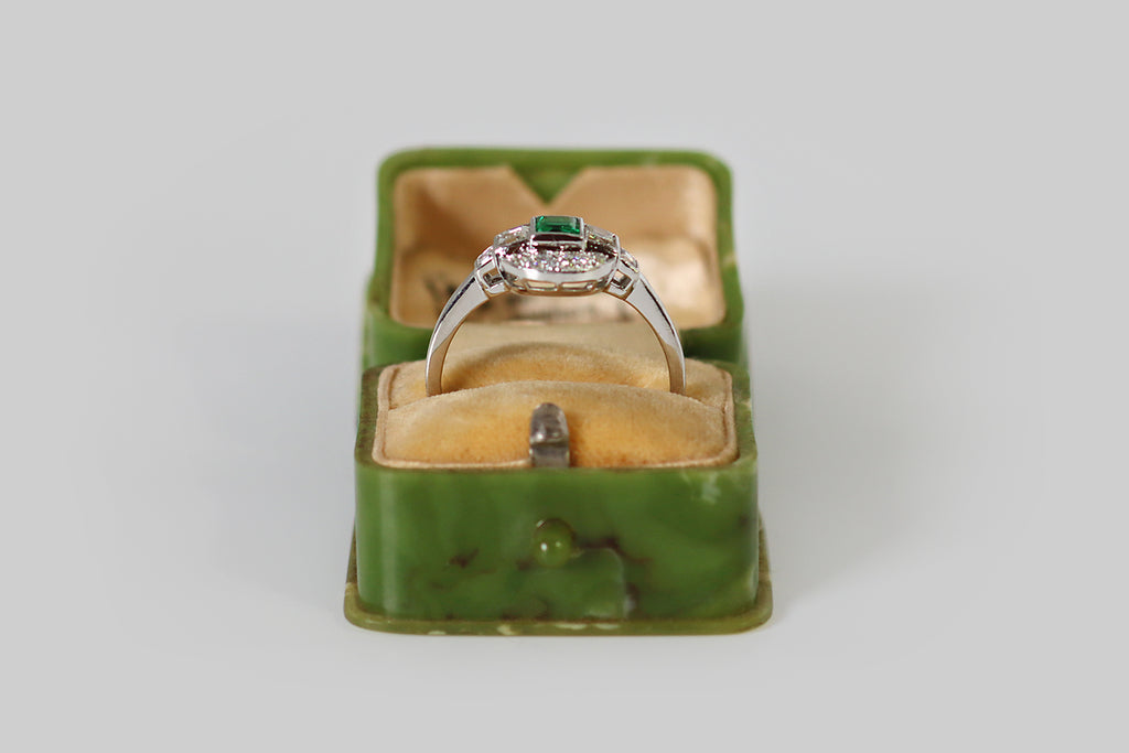 Antique Jewelry Portland, Vintage Jewelry Portland , Antique Engagement Rings | Poor Mouchette | An unusual Art Deco era engagement ring, modeled in platinum, whose primary gem is a vibrant, highly-saturated, step cut emerald. This beautiful emerald is held in a smooth, polished bezel; astride it, a pair of step-cut diamonds rest in similar bezels. These three, central gems are arranged in a linear manner, horizontally, atop the ring's circular face