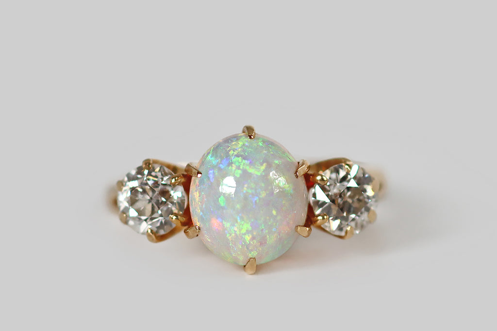 Poor Mouchette | Curated Antique Jewelry, Vintage Jewelry & Engagement Rings | A beautiful, Edwardian era three stone ring, whose primary gem is a fiery, oval, Australian opal cabochon. This crystal opal is held aloft in a crown-like, six prong mount. It has a light blue body tone, and it displays a full spectrum of shimmering colors (shades of green, blue, red, and purple), in a pinfire pattern! Astride this opal cabochon, a pair of old European cut diamonds are held in six prong mounts.