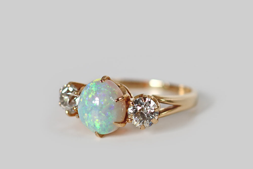 Poor Mouchette | Curated Antique Jewelry, Vintage Jewelry & Engagement Rings | A beautiful, Edwardian era three stone ring, whose primary gem is a fiery, oval, Australian opal cabochon. This crystal opal is held aloft in a crown-like, six prong mount. It has a light blue body tone, and it displays a full spectrum of shimmering colors (shades of green, blue, red, and purple), in a pinfire pattern! Astride this opal cabochon, a pair of old European cut diamonds are held in six prong mounts.