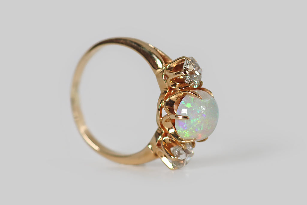 Poor Mouchette | Curated Antique Jewelry, Vintage Jewelry & Engagement Rings | A beautiful, Edwardian era three stone ring, whose primary gem is a fiery, oval, Australian opal cabochon. This crystal opal is held aloft in a crown-like, six prong mount. It has a light blue body tone, and it displays a full spectrum of shimmering colors (shades of green, blue, red, and purple), in a pinfire pattern! Astride this opal cabochon, a pair of old European cut diamonds are held in six prong mounts.