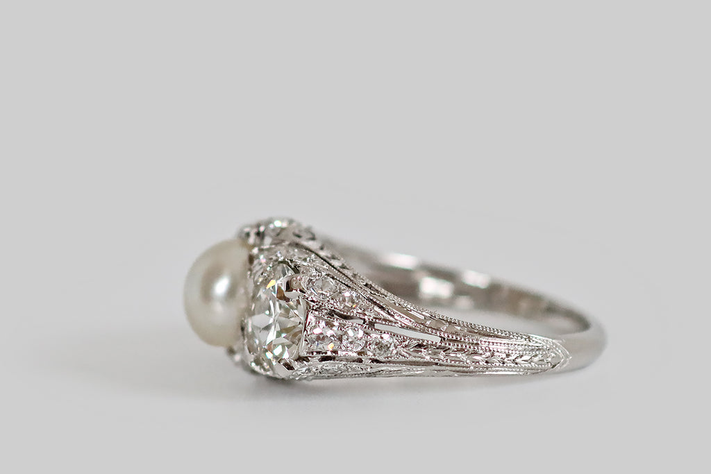Antique Jewelry Portland, Vintage Jewelry Portland , Antique Engagement Rings | Poor Mouchette | An early 20th century three stone ring, modeled in platinum, whose primary gems are a pair of old European cut diamonds (1.7 carats, together) and a luminous, natural sea pearl. These antique diamonds are held in low-profile, tab-style prongs, where they flank the ring's silvery button pearl. Framing these gems, a wave-like series of diamond-set forms that seem to "splash up" from the ring's gallery and shoulder