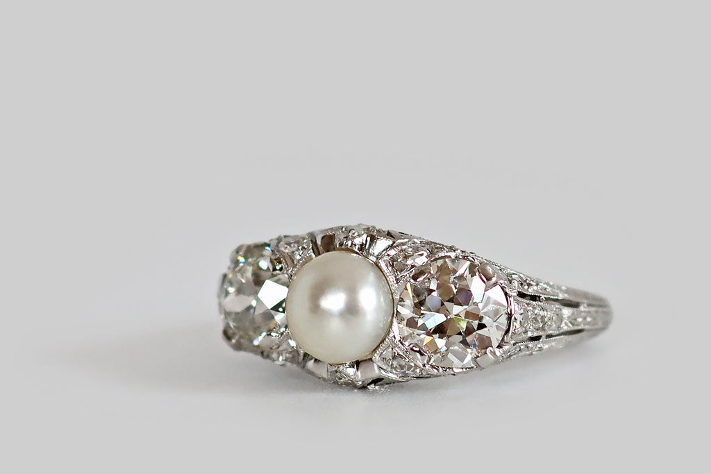 Antique Jewelry Portland, Vintage Jewelry Portland , Antique Engagement Rings | Poor Mouchette | An early 20th century three stone ring, modeled in platinum, whose primary gems are a pair of old European cut diamonds (1.7 carats, together) and a luminous, natural sea pearl. These antique diamonds are held in low-profile, tab-style prongs, where they flank the ring's silvery button pearl. Framing these gems, a wave-like series of diamond-set forms that seem to "splash up" from the ring's gallery and shoulder