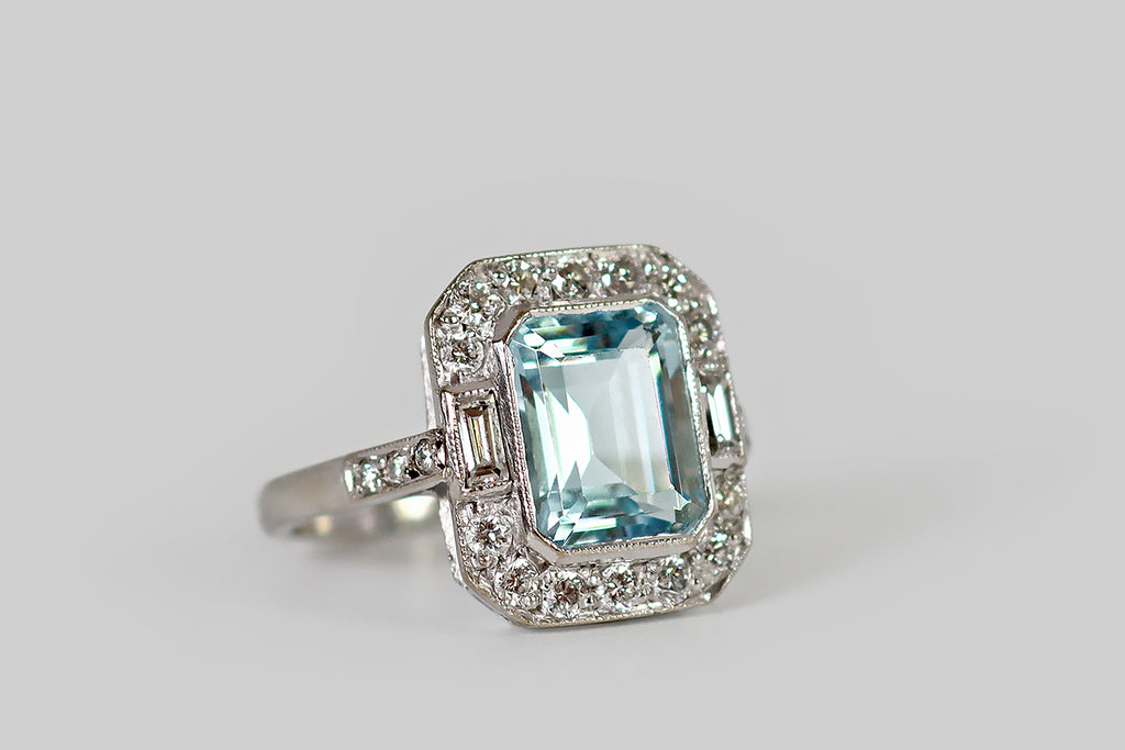Poor Mouchette | Curated Antique Jewelry, Vintage Jewelry & Engagement Rings | A late vintage, Art Deco style ring, modeled in 18k white gold, whose primary gem is a large, step-cut aquamarine (about 2.35 carats). This faceted aqua is a soft, pale blue color, inflected with subtle green undertones— it is held in a low-profile, milgrained bezel, where it is surround by a halo of fourteen, bead-set, round brilliant cut diamonds, and a pair of bezel-set baguettes.