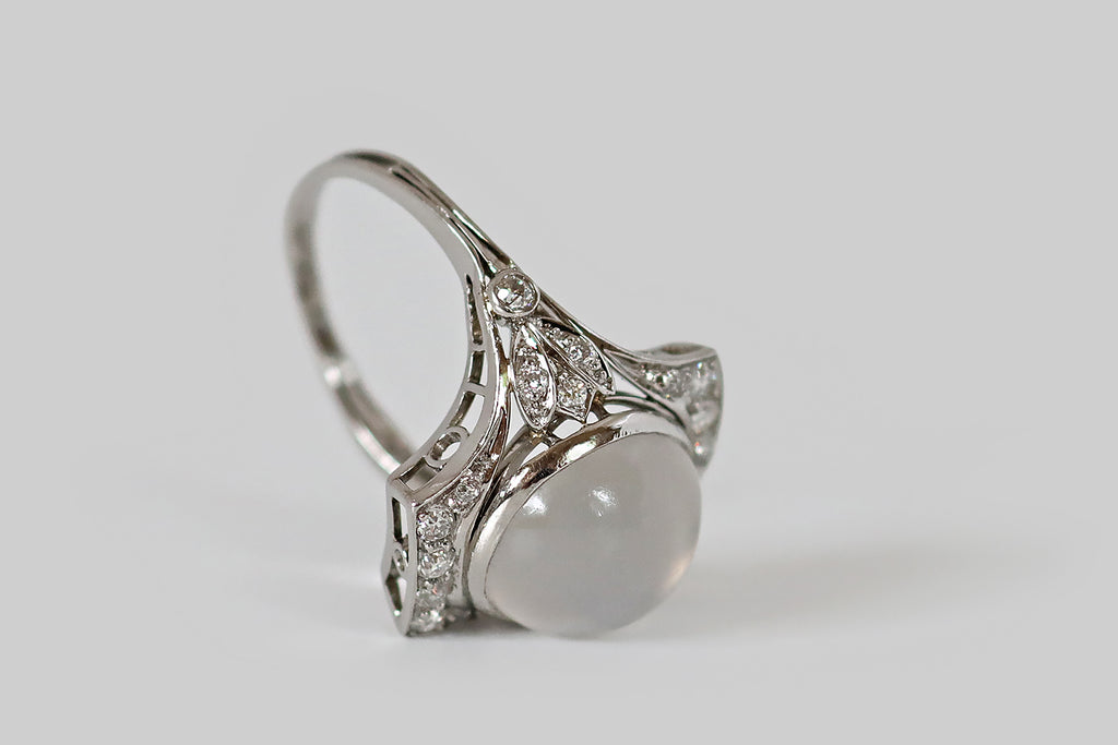 Antique Jewelry Portland, Vintage Jewelry Portland , Antique Engagement Rings | Poor Mouchette | An Art Deco era dinner ring, with subtle Egyptian flavor, modeled in platinum and featuring a large, white, cats eye moonstone. This gem has a well-defined, crisp eye that shifts across its translucent surface. The moonstone is bezel set into the ring's flared, rectangular face, which is adorned (to the north and south) with glittering, bead-set, old European cut diamonds. 