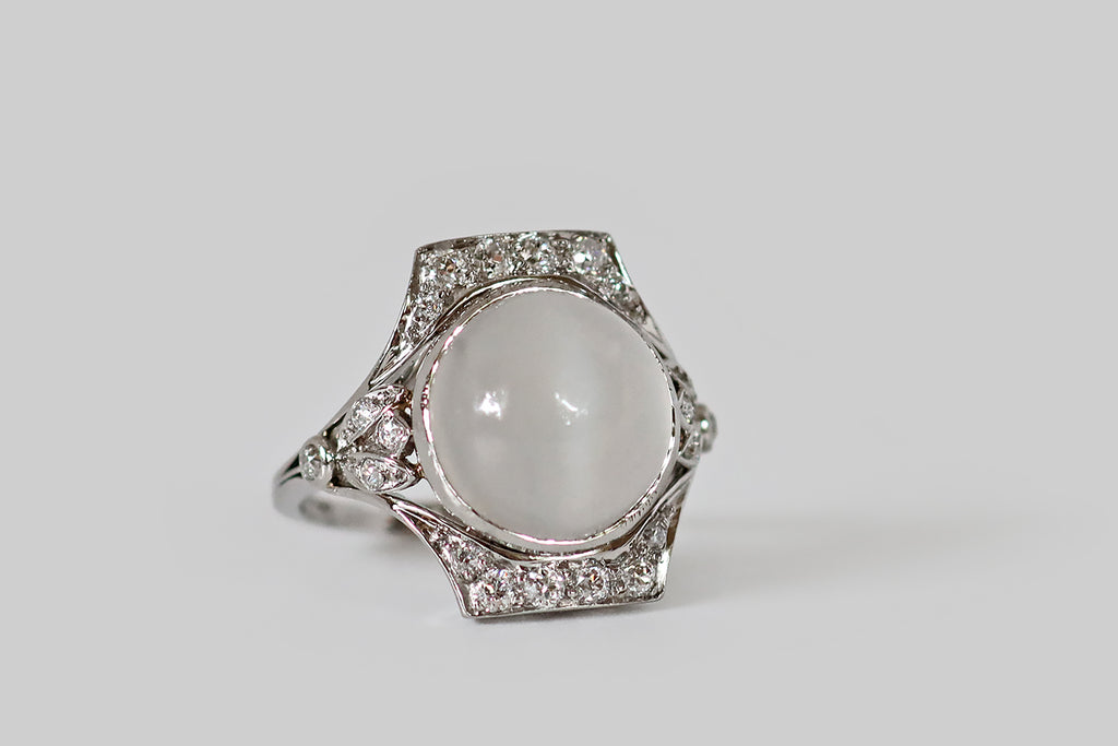 Antique Jewelry Portland, Vintage Jewelry Portland , Antique Engagement Rings | Poor Mouchette | An Art Deco era dinner ring, with subtle Egyptian flavor, modeled in platinum and featuring a large, white, cats eye moonstone. This gem has a well-defined, crisp eye that shifts across its translucent surface. The moonstone is bezel set into the ring's flared, rectangular face, which is adorned (to the north and south) with glittering, bead-set, old European cut diamonds. 