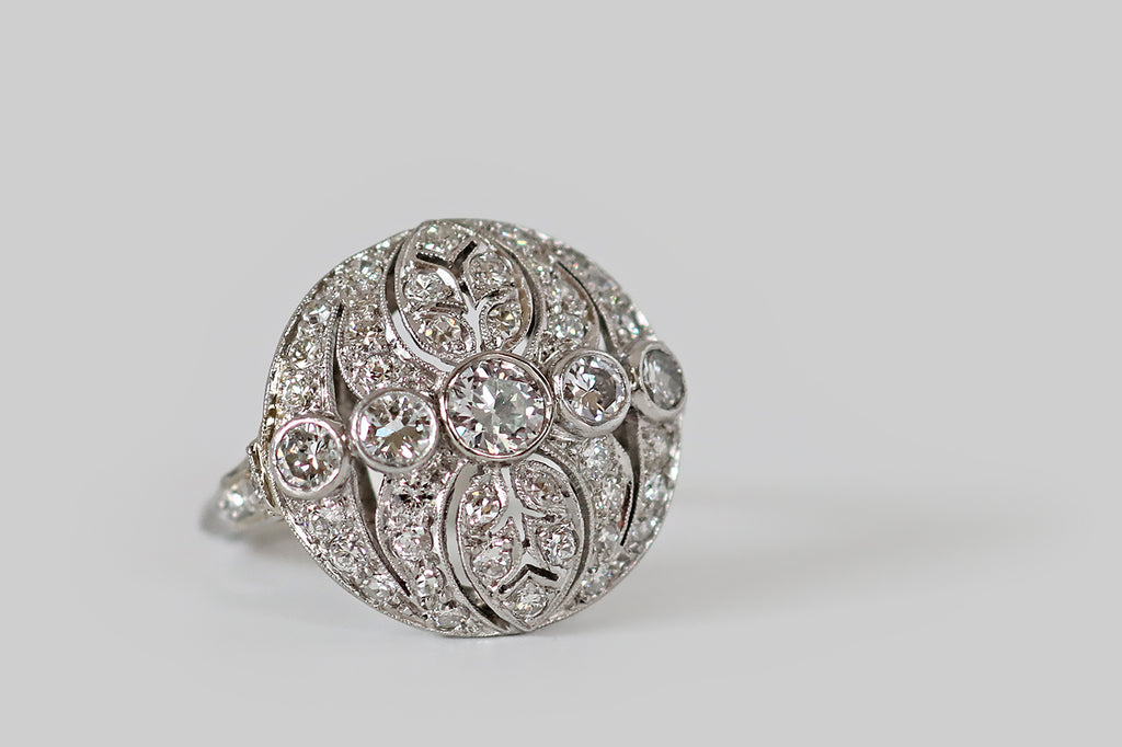 Antique Jewelry Portland, Vintage Jewelry Portland , Antique Engagement Rings | Poor Mouchette | A circular, Art Deco era diamond cluster ring, modeled in platinum, centering on a series of five bezel-set diamonds (a primary Old European cut diamond and four transitionals) which are arranged horizontally across its intricate, openwork face. This openwork features a pair of pierced, ovate leaves (reminiscent of philodendron), flanked by concentric crescent shapes that bow toward the center