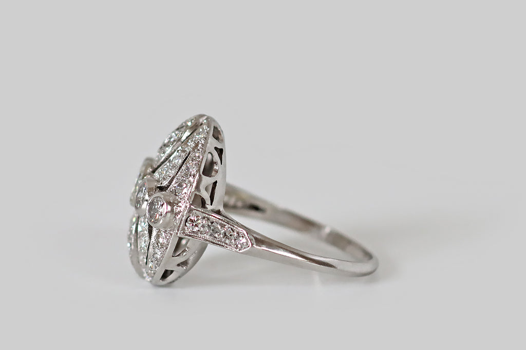 Antique Jewelry Portland, Vintage Jewelry Portland , Antique Engagement Rings | Poor Mouchette | A circular, Art Deco era diamond cluster ring, modeled in platinum, centering on a series of five bezel-set diamonds (a primary Old European cut diamond and four transitionals) which are arranged horizontally across its intricate, openwork face. This openwork features a pair of pierced, ovate leaves (reminiscent of philodendron), flanked by concentric crescent shapes that bow toward the center