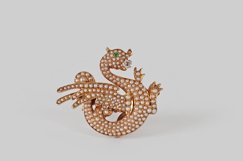Antique Jewelry Portland, Vintage Jewelry Portland , Antique Engagement Rings | Poor Mouchette | An Art Nouveau era brooch, modeled as an adorable wyvern (dragon) in 14k yellow gold. Our beautiful beast is dimensional, highly stylized, and she is covered with a multitude of tiny natural pearls that represent her scales! She is winged, with two legs, a curling tail, and a vibrant green demantoid garnet eye. She hold an old European cut diamond in her mouth— a treasure that traditionally represents wisdom