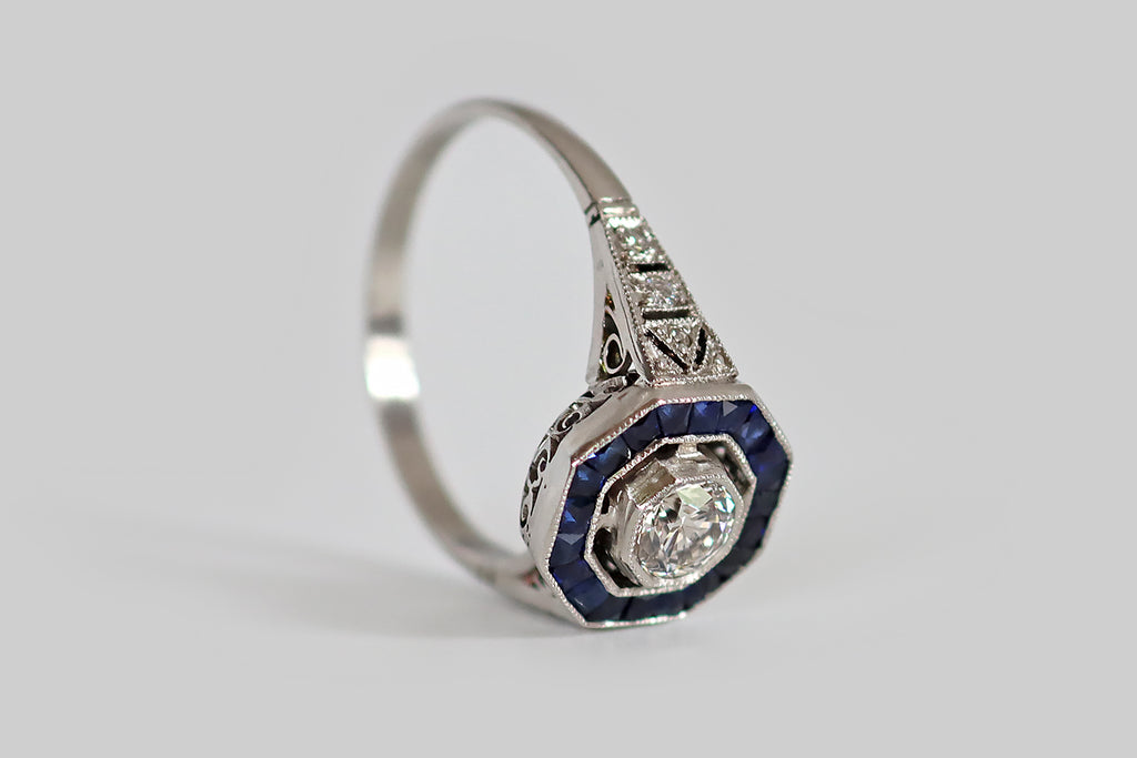 Antique Jewelry Portland, Vintage Jewelry Portland , Antique Engagement Rings | Poor Mouchette | An Art Deco era hexagonal halo ring, modeled in platinum, whose primary gem is a 1/3 carat transitional cut diamond (G-H, VS). This ring has an elegant, oft-imitated, iconic form— its bezel-set diamond floats at center, surrounded by a border of French cut, blue sapphires, that are calibre set into their own milgrained channel. Its diamond-set shoulders are decoratively pierced in the manner of epaulettes