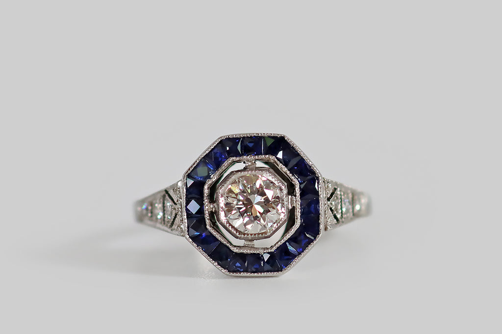Antique Jewelry Portland, Vintage Jewelry Portland , Antique Engagement Rings | Poor Mouchette | An Art Deco era hexagonal halo ring, modeled in platinum, whose primary gem is a 1/3 carat transitional cut diamond (G-H, VS). This ring has an elegant, oft-imitated, iconic form— its bezel-set diamond floats at center, surrounded by a border of French cut, blue sapphires, that are calibre set into their own milgrained channel. Its diamond-set shoulders are decoratively pierced in the manner of epaulettes