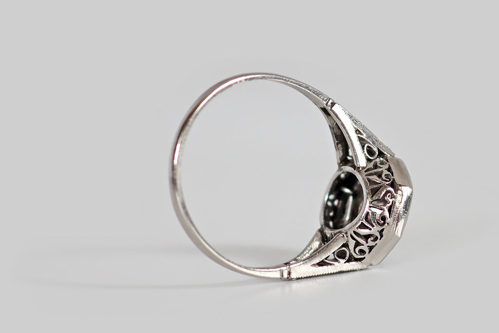 Antique Jewelry Portland, Vintage Jewelry Portland , Antique Engagement Rings | Poor Mouchette | An Art Deco era hexagonal halo ring, modeled in platinum, whose primary gem is a 1/3 carat transitional cut diamond (G-H, VS). This ring has an elegant, oft-imitated, iconic form— its bezel-set diamond floats at center, surrounded by a border of French cut, blue sapphires, that are calibre set into their own milgrained channel. Its diamond-set shoulders are decoratively pierced in the manner of epaulettes