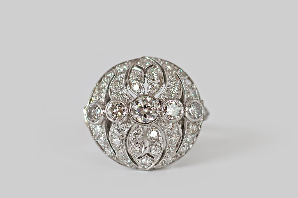 Antique Jewelry Portland, Vintage Jewelry Portland , Antique Engagement Rings | Poor Mouchette | A circular, Art Deco era diamond cluster ring, modeled in platinum, centering on a series of five bezel-set diamonds (a primary Old European cut diamond and four transitionals) which are arranged horizontally across its intricate, openwork face. This openwork features a pair of pierced, ovate leaves (reminiscent of philodendron), flanked by concentric crescent shapes that bow toward the center