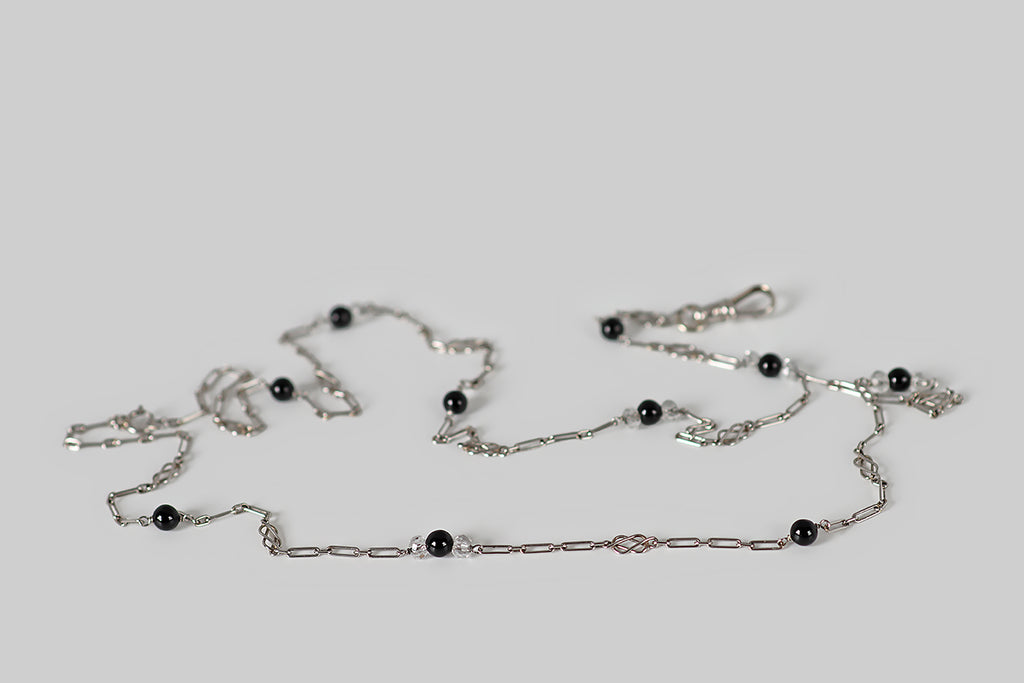 Antique Jewelry Portland, Vintage Jewelry Portland , Antique Engagement Rings | Poor Mouchette | A delicate Art Deco era lariat, modeled in platinum, whose lengths of elongated oval cable links are punctuated by a series of spacers— single onyx beads, trios of onyx/faceted-crystal, and tiny, hand-formed love knots! This airy platinum chain is fitted with a small, white gold dog clip that drops from the end of its "y." This little dog clip can hold multiple charms or a pendant.