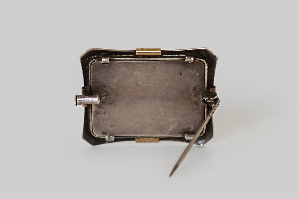 Antique Jewelry Portland, Vintage Jewelry Portland , Antique Engagement Rings | Poor Mouchette | An Arts &amp; Crafts era brooch, imagined as a picture frame, with a 14k gold face, and a sterling silver back. This elegant rectangular frame is gathered at the north, south, east, and west, where it is embellished with notched, clip-like forms. The frames clipped corners are lightly dapped, to add dimensionality. Secure beneath the frame's softly-beveled crystal, a hand-painted watercolor miniature