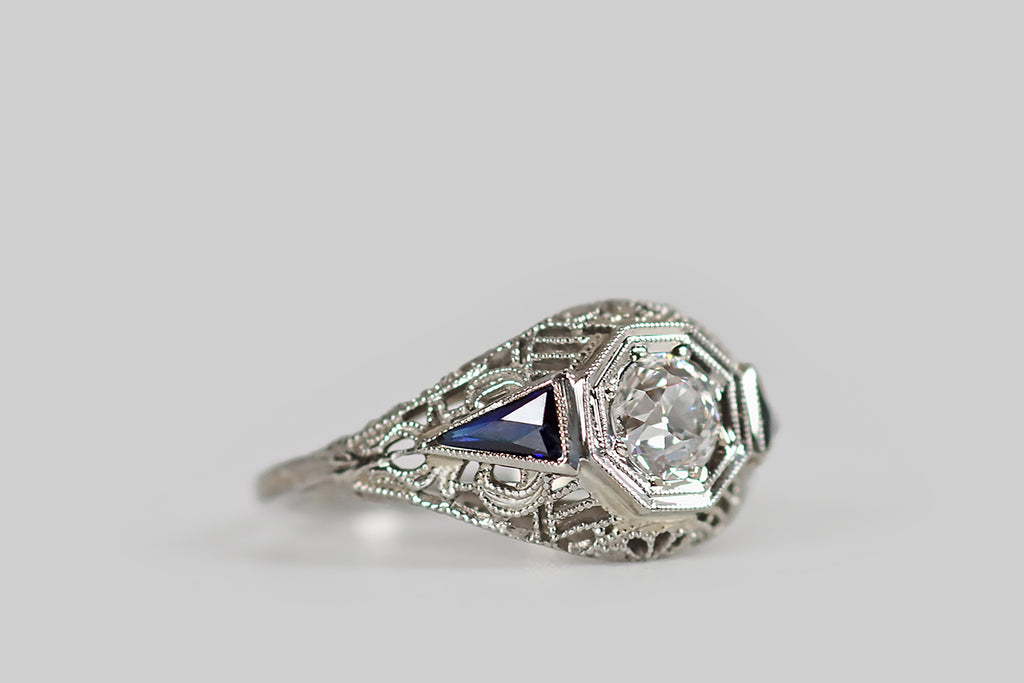 Antique Jewelry Portland, Vintage Jewelry Portland , Antique Engagement Rings | Poor Mouchette | A classic, Art Deco era, filigree engagement ring, modeled in 18k white gold and set with a chunky .52 carat old European cut diamond (F/G, SI1). This sparkling, white diamond is bead-set into an octagonal seat at the center of the ring face, which is low-profile and softly domed. It is flanked by a pair of deep blue, elongated, pennant-shaped, created sapphires.