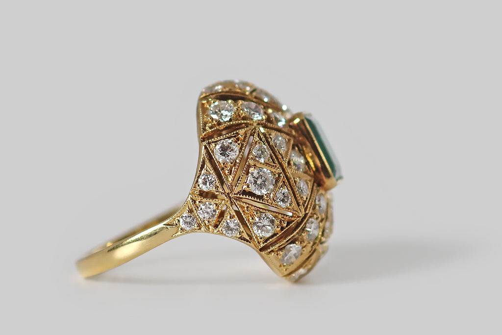 Antique Jewelry Portland, Vintage Jewelry Portland , Antique Engagement Rings | Poor Mouchette | A glamorous, mid-twentieth-century ring, modeled in 18k yellow gold, whose cushion-shaped face is encrusted with an ornate arrangement of gemstones— many, sparkling round brilliant cut diamonds, surrounding a central, rich-green, step-cut emerald.&nbsp;This ring has a soft, bombe-like profile. The emerald rests at the center of the ring face