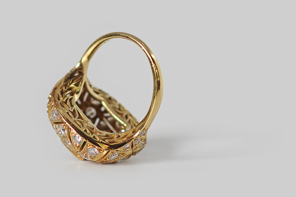 Antique Jewelry Portland, Vintage Jewelry Portland , Antique Engagement Rings | Poor Mouchette | A glamorous, mid-twentieth-century ring, modeled in 18k yellow gold, whose cushion-shaped face is encrusted with an ornate arrangement of gemstones— many, sparkling round brilliant cut diamonds, surrounding a central, rich-green, step-cut emerald.&nbsp;This ring has a soft, bombe-like profile. The emerald rests at the center of the ring face