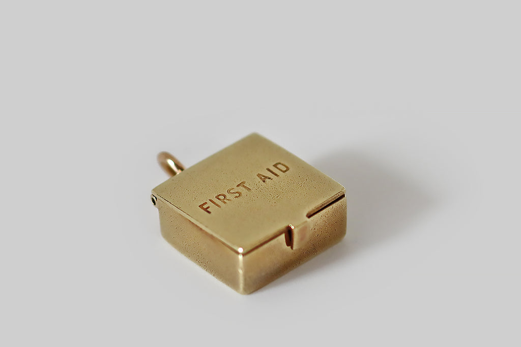 Antique Jewelry Portland, Vintage Jewelry Portland , Antique Engagement Rings | Poor Mouchette | A dainty, vintage charm, modeled as a first aid kit, in 14k yellow gold. This beautifully made first aid box opens to reveal a group of tiny medical supplies— a roll of "gauze," a bottle of "pills," etc., all enameled in red, blue, and white. This is marked 14k for purity and for the maker Sloan & Co. It hangs from an oval jump ring, that is attached to the medical box's handle.