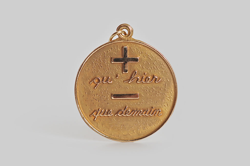 Poor Mouchette | Curated Antique Jewelry, Vintage Jewelry & Engagement Rings | A large, circular pendant, modeled in 14k yellow gold, that bears the well-known sentiment "+ qu'hier - que demain": more than yesterday, less than tomorrow. This phrase is rendered in polished relief, atop the pendant's textured, lightly-patinead face. We love this for its bold, graphic quality and sweet sentiment!