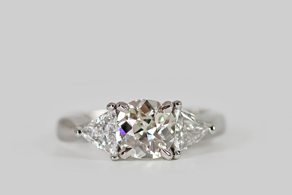Antique Jewelry Portland, Vintage Jewelry Portland , Antique Engagement Rings | Poor Mouchette | A striking, 1940s, three-stone engagement ring, modeled in platinum, whose central, old mine cushion cut diamond is flanked by a pair of antique triangle cuts. The old mine cut diamond is vibrant, with flashing, slightly-irregular facets, a small table, and a large open culet. The antique triangles are bright and sparkly, with many, slender, ray-like facets and tiny, open culets.
