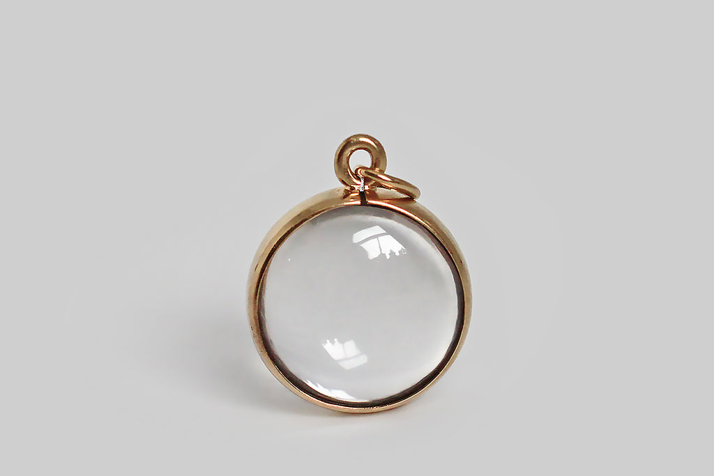 Antique Jewelry Portland, Vintage Jewelry Portland , Antique Engagement Rings | Poor Mouchette | A dainty, mid 20th century charm, whose broad 14k yellow gold bezel frames an optically clear, two-domed, crystal cabochon. This little charm is strikingly minimal— its transparent crystal reads like a drop of water, suspended inside a gold ring. It has a serene visual quality when worn alone on a slender chain. This charm was probably intended as a small magnifying glass.