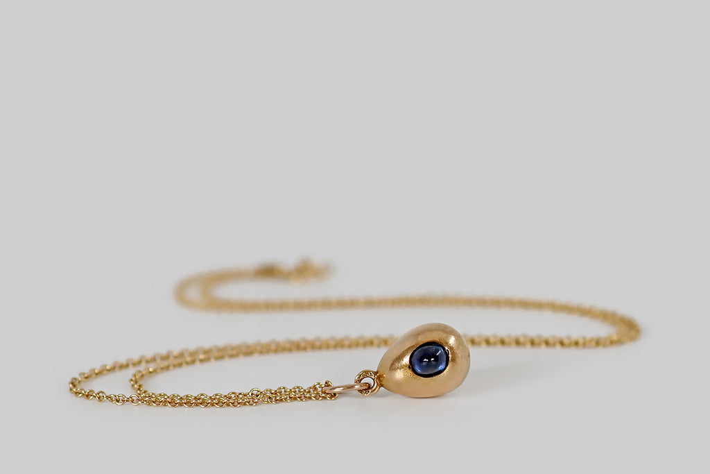 Antique Jewelry Portland, Vintage Jewelry Portland , Antique Engagement Rings | Poor Mouchette | A miniature, imperial Russian egg pendant, modeled in 14k yellow gold, whose single embellishment is a peaked, oval, medium-blue, natural, sapphire cabochon. This glowing, sugarloaf sapphire nests in the face of our little egg, where it is secured by the low profile edge of its seat, and a pair of graved beads.