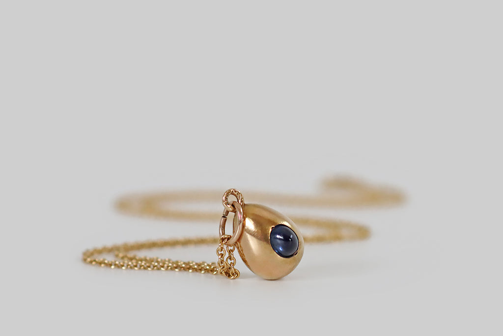 Antique Jewelry Portland, Vintage Jewelry Portland , Antique Engagement Rings | Poor Mouchette | A miniature, imperial Russian egg pendant, modeled in 14k yellow gold, whose single embellishment is a peaked, oval, medium-blue, natural, sapphire cabochon. This glowing, sugarloaf sapphire nests in the face of our little egg, where it is secured by the low profile edge of its seat, and a pair of graved beads.
