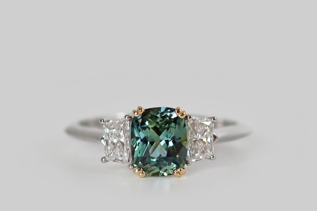Antique Jewelry Portland, Vintage Jewelry Portland , Antique Engagement Rings | Poor Mouchette | An elegant, handmade, three stone ring, modeled in platinum and 18k gold, whose primary gem is a natural, untreated, teal-colored Sri Lankan sapphire (1.37 carats). This elongated, cushion cut sapphire is held in a yellow gold, split-prong basket, where it is flanked by a pair of white, prong-set, trapezoid diamonds (F/G, VS1). 