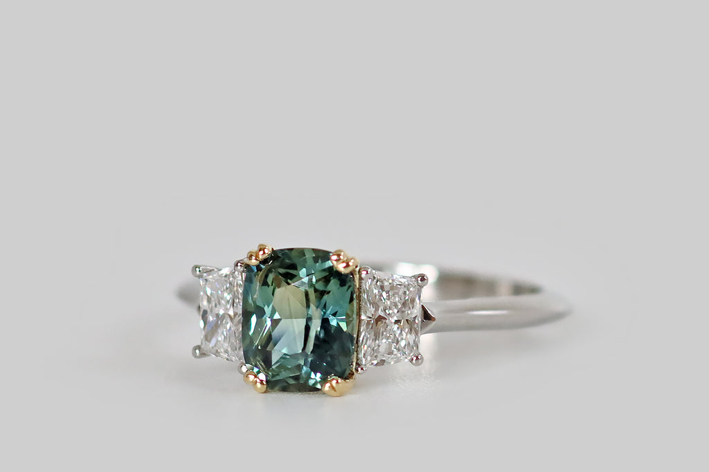 Antique Jewelry Portland, Vintage Jewelry Portland , Antique Engagement Rings | Poor Mouchette | An elegant, handmade, three stone ring, modeled in platinum and 18k gold, whose primary gem is a natural, untreated, teal-colored Sri Lankan sapphire (1.37 carats). This elongated, cushion cut sapphire is held in a yellow gold, split-prong basket, where it is flanked by a pair of white, prong-set, trapezoid diamonds (F/G, VS1). 
