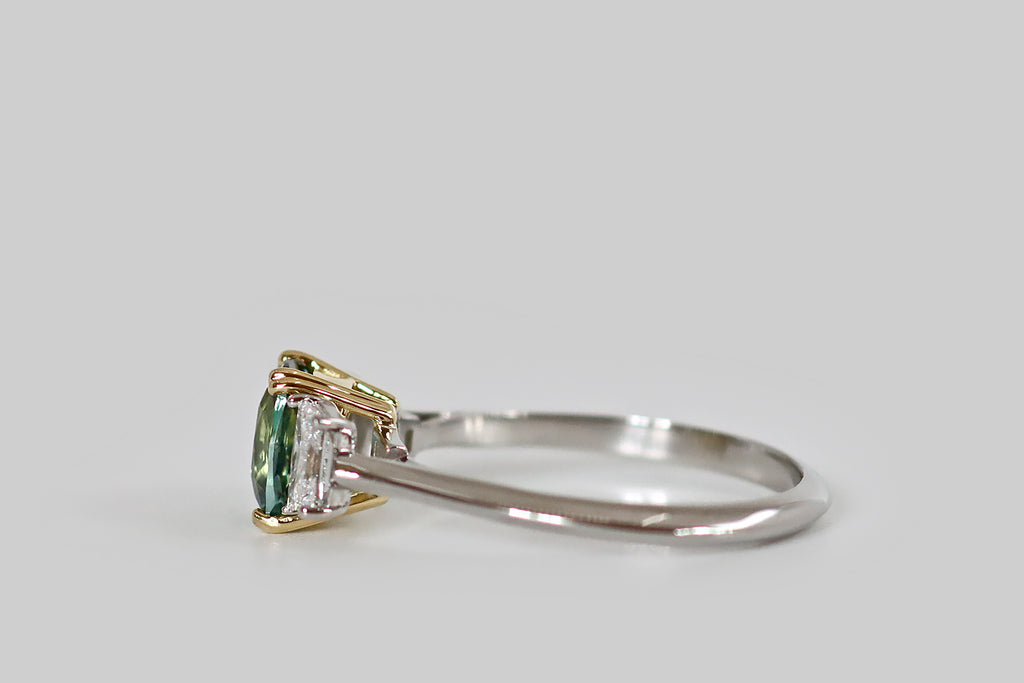 Antique Jewelry Portland, Vintage Jewelry Portland , Antique Engagement Rings | Poor Mouchette | An elegant, handmade, three stone ring, modeled in platinum and 18k gold, whose primary gem is a natural, untreated, teal-colored Sri Lankan sapphire (1.37 carats). This elongated, cushion cut sapphire is held in a yellow gold, split-prong basket, where it is flanked by a pair of white, prong-set, trapezoid diamonds (F/G, VS1). 