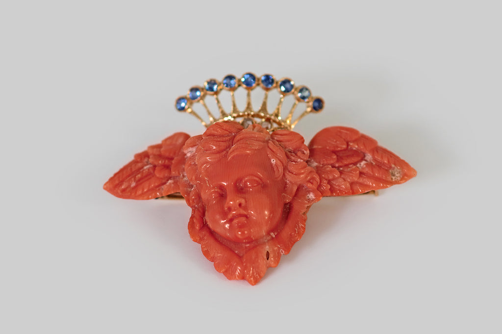 Poor Mouchette | Curated Antique Jewelry, Vintage Jewelry & Engagement Rings | Portland, Oregon | A large, Victorian era, carved coral brooch, whose subject is a cherub's head, resting between a pair of feathered wings. An ornate, celestial crown, modeled in 18k yellow gold, hovers behind the head of our resting angel. This crown's nine, tapering rays terminate in bezels that hold natural, old cut, medium blue sapphires; below these rays, a band decorated with five rose cut diamonds, set in diamond-shaped 