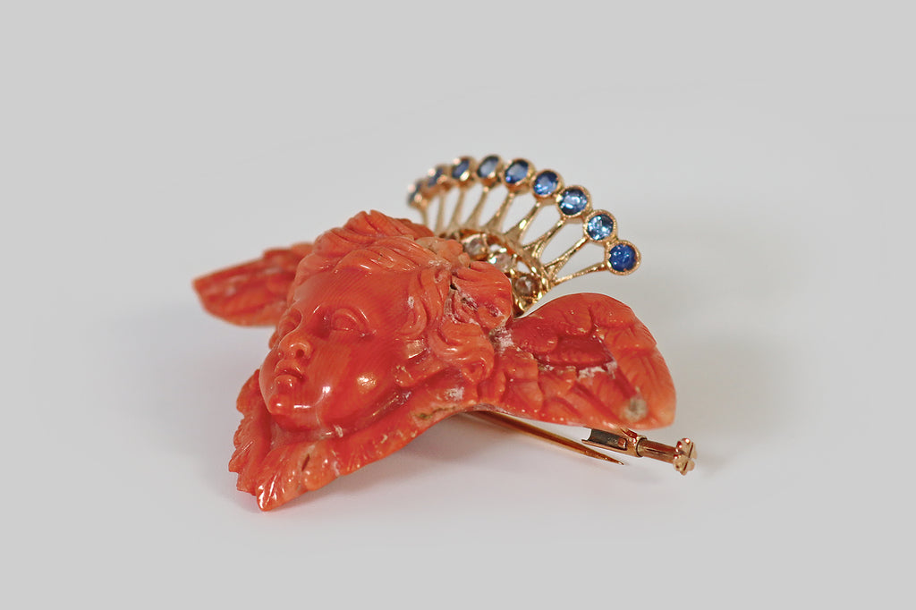 Poor Mouchette | Curated Antique Jewelry, Vintage Jewelry & Engagement Rings | Portland, Oregon | A large, Victorian era, carved coral brooch, whose subject is a cherub's head, resting between a pair of feathered wings. An ornate, celestial crown, modeled in 18k yellow gold, hovers behind the head of our resting angel. This crown's nine, tapering rays terminate in bezels that hold natural, old cut, medium blue sapphires; below these rays, a band decorated with five rose cut diamonds, set in diamond-shaped 