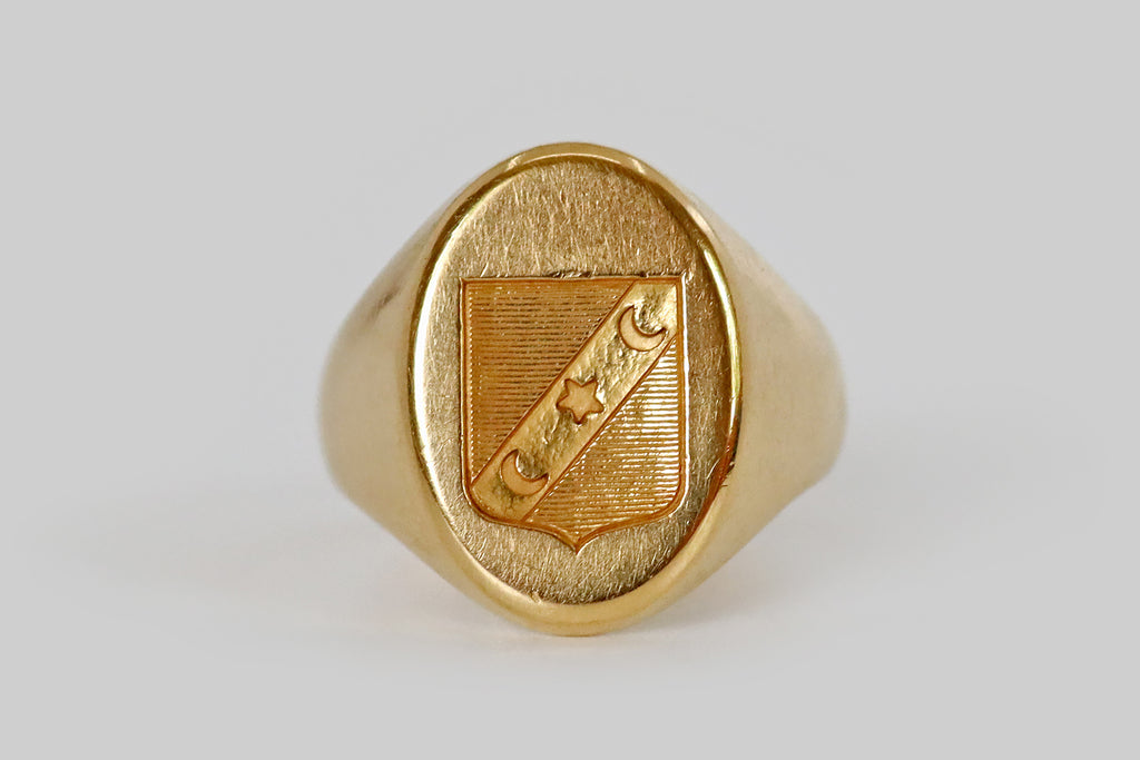 Poor Mouchette | Curated Antique Jewelry, Vintage Jewelry & Engagement Rings | A large scale, Victorian era signet ring, modeled in 18k yello gold, whose long, oval face is emblazoned with a heraldic shield. Atop a field of fine horizontal stripes, a bend, decorated with two crescent moons and a star, crosses the face of this shield. This weighty antique ring has the classic signet profile