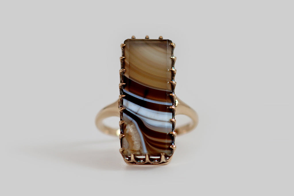 Antique Jewelry Portland, Vintage Jewelry Portland , Antique Engagement Rings | Poor Mouchette | An unusual Victorian era ring, modeled in 14k rosy-yellow gold, whose gem is an extra long, rectangular, polished agate slab with black, brown, and white banding. This linear figuring sweeps across the agate— it is lightest, and most translucent at the top, becoming richer, darker, and more saturated toward the bottom of the stone. A pocket a druzy crystals