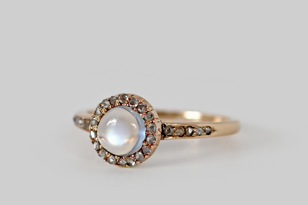 Antique Jewelry Portland, Vintage Jewelry Portland , Antique Engagement Rings | Poor Mouchette | An early twentieth century ring, modeled in 14k rose gold, whose primary gem is a round, blue-flash moonstone cabochon. This moonstone is held in a nest of eighteen bead-like prongs, where it is surrounded by a halo of eighteen silvery rose cut diamonds. These tiny rose cuts share prongs with the moonstone, and they are further secured by the ring's crimped outer bezel. 