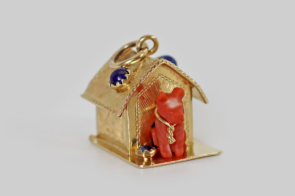 Antique Jewelry Portland, Vintage Jewelry Portland , Antique Engagement Rings | Poor Mouchette |A large, mid twentieth century charm, fabricated in 18k yellow gold, resembling a classic doghouse with a pup inside! Our faithful doggo was hand-carved from vibrant Sardinian coral, and the carver has positioned a naturally-occurring white spot in the middle of his forehead. He sits inside the doorway of his gold house, wearing a twisted gold tie out— beside him, a water bowl (a lapis lazuli cabochon.