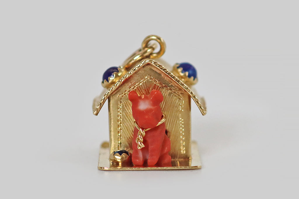 Antique Jewelry Portland, Vintage Jewelry Portland , Antique Engagement Rings | Poor Mouchette |A large, mid twentieth century charm, fabricated in 18k yellow gold, resembling a classic doghouse with a pup inside! Our faithful doggo was hand-carved from vibrant Sardinian coral, and the carver has positioned a naturally-occurring white spot in the middle of his forehead. He sits inside the doorway of his gold house, wearing a twisted gold tie out— beside him, a water bowl (a lapis lazuli cabochon.