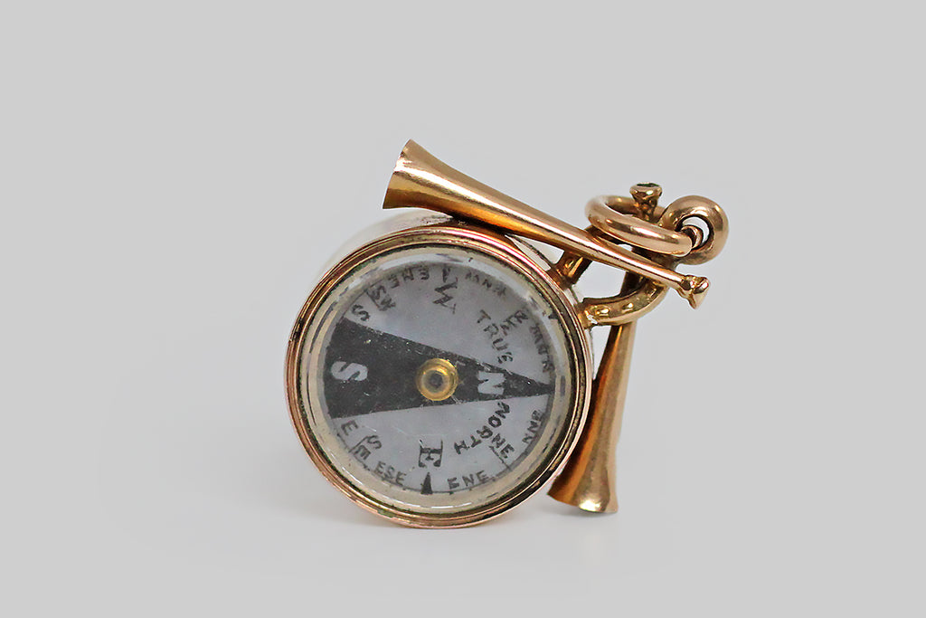 An unusual, Victorian-era, working, compass watch fob, modeled in 18k rosy yellow gold, and crowned with a sleek pair of hunt horns and a small horseshoe. The compass face is silvery, with black markings to indicate the cardinal and inter-cardinal directions; the words "true north" are printed alongside the needle that indicates North. 