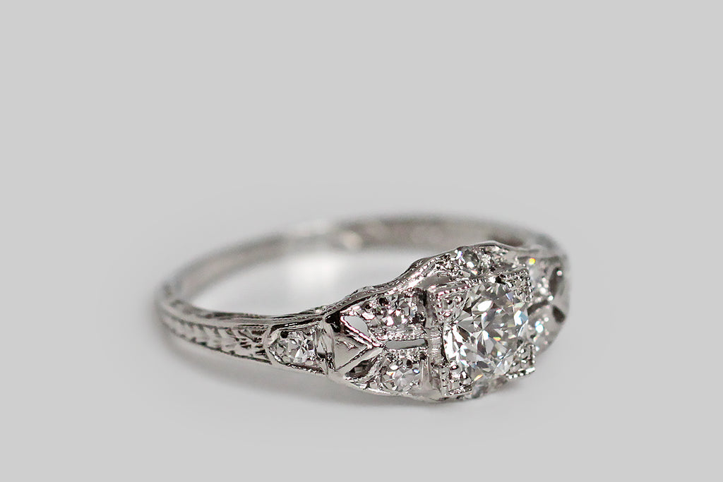Poor Mouchette | Curated Antique Jewelry, Vintage Jewelry & Engagement Rings | Portland, Oregon | An elegant, 1930s engagement ring, modeled in platinum, whose primary gem is a 1/2 carat old European cut diamond (F/G, VS). This sparkling, hand-cut sweetheart is mounted in a low-profile, four-tab basket setting, whose "picture corner" style tabs are embellished with fine milgrain, and clover-like trios of beads. The ring head rests atop the delicate, shapely face, surrounded by pave-set, single-cut diamonds