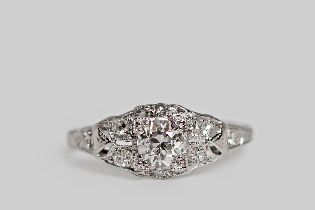 Poor Mouchette | Curated Antique Jewelry, Vintage Jewelry & Engagement Rings | Portland, Oregon | An elegant, 1930s engagement ring, modeled in platinum, whose primary gem is a 1/2 carat old European cut diamond (F/G, VS). This sparkling, hand-cut sweetheart is mounted in a low-profile, four-tab basket setting, whose "picture corner" style tabs are embellished with fine milgrain, and clover-like trios of beads. The ring head rests atop the delicate, shapely face, surrounded by pave-set, single-cut diamonds