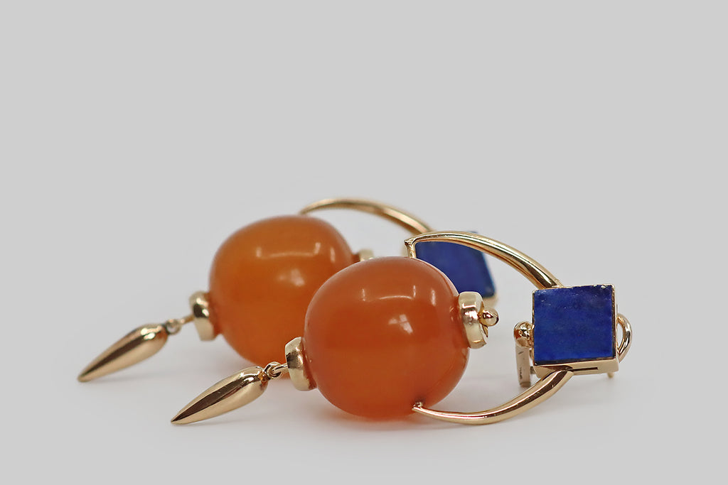 Poor Mouchette | Curated Antique Jewelry, Vintage Jewelry & Engagement Rings | Portland, Oregon | A bold, graphic pair of late-vintage earrings, modeled in 14k yellow gold, whose pendant elements are large, plump amber (or copal) beads, that swing back and forth on gold frames. These tong-like frames hang below the earring tops, which are set with pyrite-flecked slabs of bright blue lapis lazuli— the square tops also tip backward and forward on the frame, as if hinged.